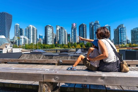 Tourist mother pointing at the skyline of vancouver while sitting with her son on a wooden bench at