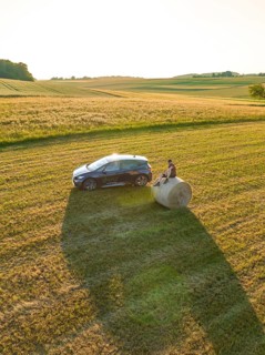 Car with person on a hay bale in a wide field at sunset, deer e-car sharing, Cupra electric car,