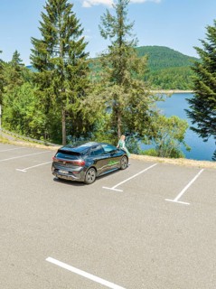 A car is parked in a parking lot with a view of a lake and surrounded by forest and mountains, deer