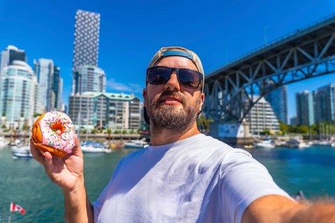 Tourist taking a selfie while holding a donut with burrard bridge and the cityscape of vancouver in