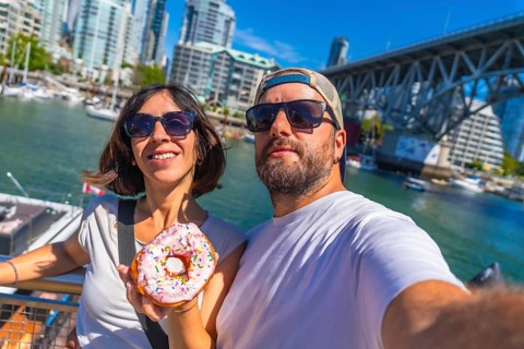 Happy tourists taking a selfie while holding a donut with sprinkles in front of the cityscape and