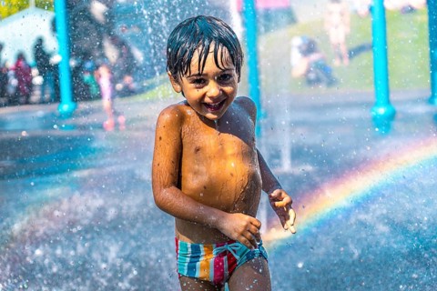Happy child running and playing under splash pad jets with rainbow in the background on a hot
