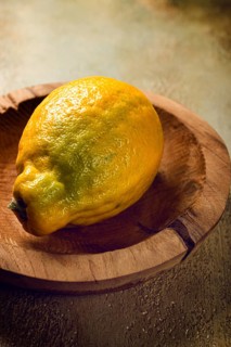 A vibrant lemon with a textured skin sits on a handcrafted wooden plate, illuminated by gentle