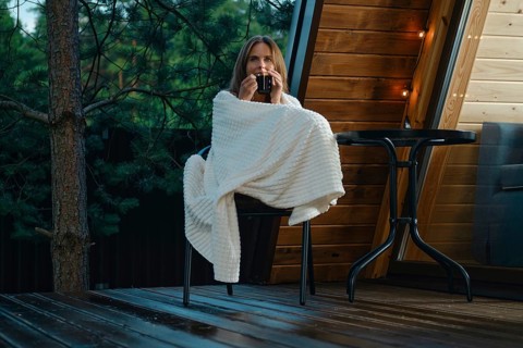 A woman relaxes on a chair while wrapped in a blanket. She holds a drink and enjoys fresh air after