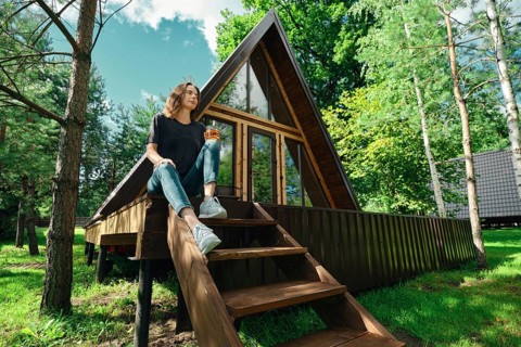 A woman sits on the steps of a wooden cabin surrounded by trees. She holds a drink and looks
