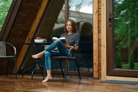Barefoot woman sits on a chair on a wet wooden deck of tiny house after the rain. She is reading a