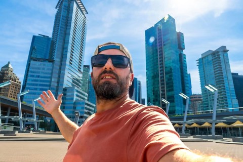 Bearded tourist wearing sunglasses and baseball cap takes selfie with outstretched arm in front of