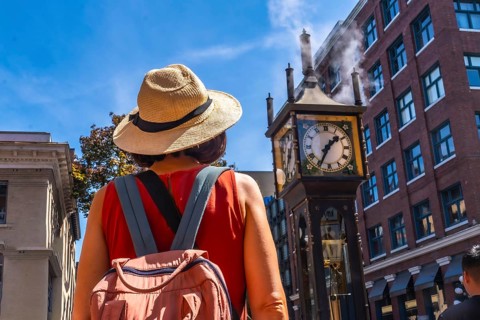 Female tourist wearing straw hat and backpack looking at the famous steam clock in gastown historic