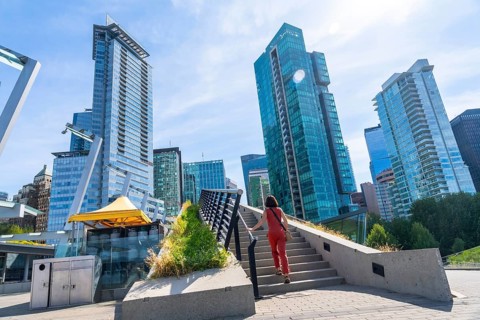 Woman is climbing the stairs of a pedestrian overpass with modern skyscrapers and a clear blue sky