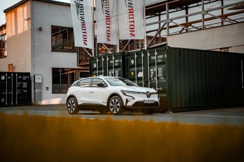 White modern SUV in front of an industrial building with containers and banners, Deer E-Carsharing,