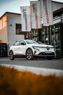 White electric car in front of a Motorworld building with flags and containers, Deer e-Carsharing,