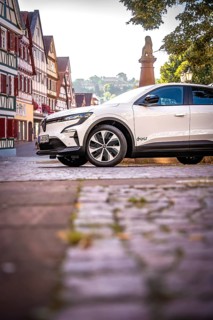 White car on cobblestone road surrounded by half-timbered houses and trees, Deer E-Carsharing,