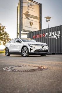 A white electric car is parked in front of a Motorworld sign on a sidewalk, Deer E- Carsharing,
