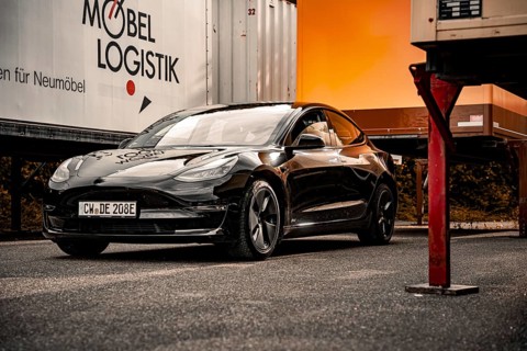 Black car in front of furniture trailers in bright sunlight on a road surface, Deer E-Carsharing,