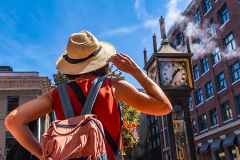 Young woman with backpack and straw hat looking at the famous steam clock in gastown, vancouver,