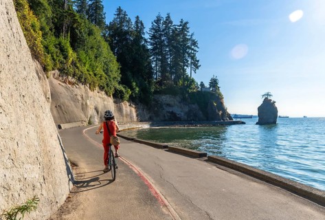 Tourist wearing helmet and red clothes enjoys cycling in stanley park seawall with siwash rock and
