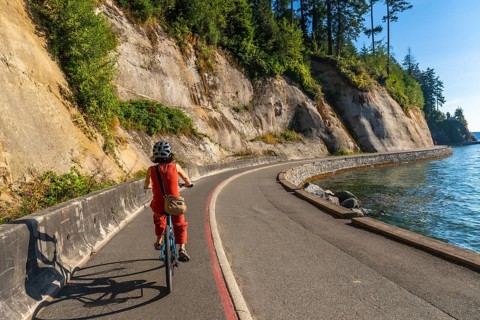 Tourist wearing helmet and carrying bag is riding bicycle along stanley park seawall in vancouver,