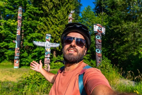 Cyclist wearing a helmet and sunglasses takes a selfie with totem poles at brockton point in
