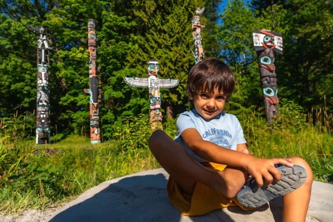 Young boy sitting on a rock, tying his shoe in front of vibrant totem poles at brockton point in