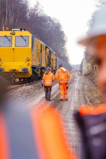 Two workers in orange on a railroad construction site next to a large yellow vehicle, tamping