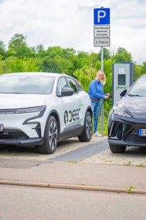 Woman at a charging station between two electric cars in a green parking lot, Deer E-Carsharing,