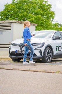 Woman standing on the street with a white electric car with charging cable, Deer E-Carsharing,