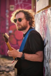 Hipster street musician in black playing electric guitar in the street on sunset leaning on a wall.