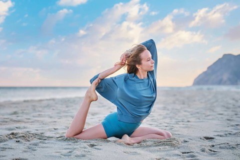 A woman practices Eka Pada Rajakapotasana on the sand during sunset, focusing on her yoga pose with