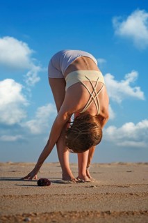 A woman performs Uttanasana on a sandy beach during sunset while clouds fill the sky. She leans