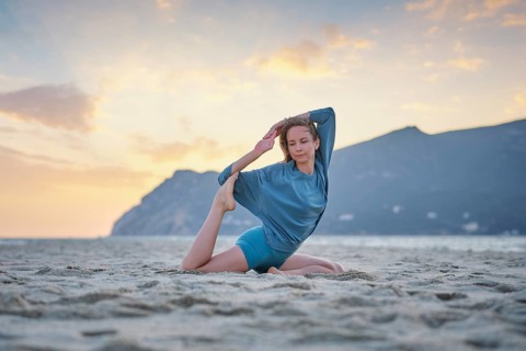 A woman performs Eka Pada Rajakapotasana on a sandy beach at sunset. The warm sky adds to the