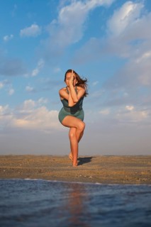 A woman stands on a sandy beach performing Eagle Pose with wrapped legs and crossed arms during