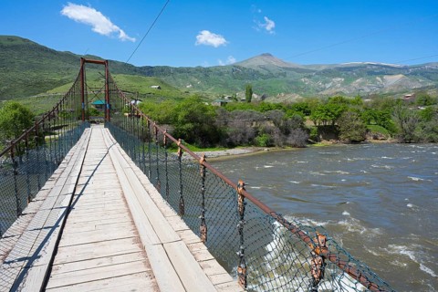 View of a swinging suspension bridge spanning a river, with green hills and a mountain in the