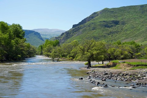 A wide river flows through lush green countryside surrounded by high hills and clear skies, Kura