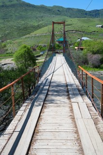 A wooden suspension bridge stretches across a river with a green landscape, Kura River near