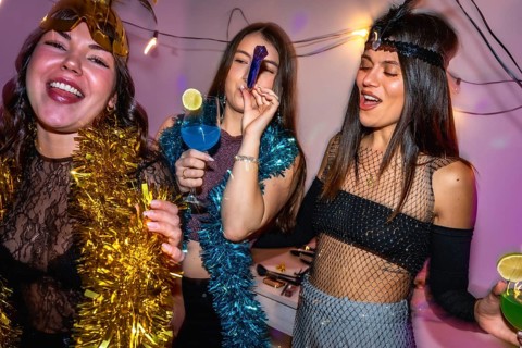 Young women in vibrant costumes laugh and clink cocktails at a festive new year's eve party