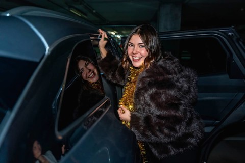 Young woman in a fur coat with gold tinsel smiles as she steps out of a car in a parking lot at