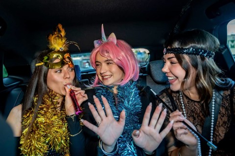 Three young women in festive costumes ride in a car, wearing tinsel and makeup, laughing and