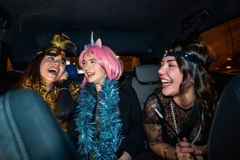 Young women sitting in the back seat of a car, wearing festive costumes and garlands, sharing a