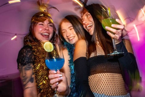 Three happy young adult women laughing while holding colorful cocktails, celebrating together with