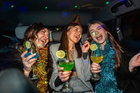 Three friends dressed up in party hats and tinsel garlands laughing and toasting colorful cocktails