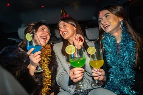 Three happy young women traveling in a car, laughing and toasting colorful cocktails together,