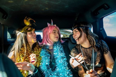 Three young women laugh and celebrate in a car during a colorful costume party, wearing masks, wigs