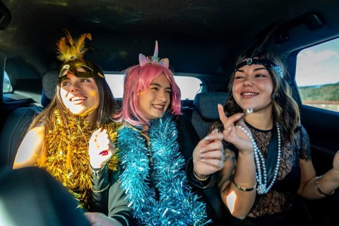 Three young women laughing and celebrating in a car back seat, wearing colorful tinsel boas and
