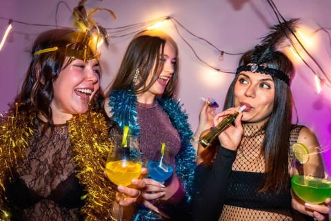 Three diverse young women in festive costumes laughing and toasting with colorful cocktails and