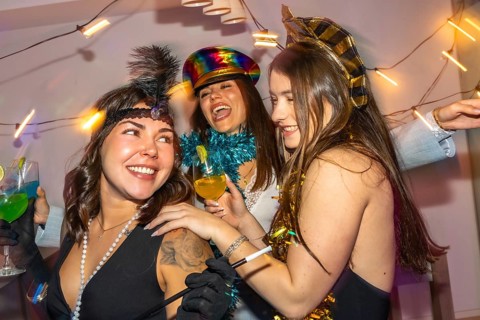 Young women in festive costumes laugh and toast cocktails at a colorful new year's eve home party,