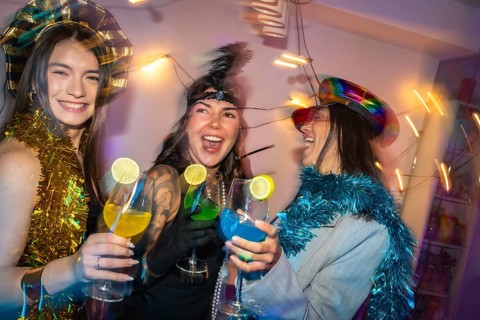 Diverse women in festive party hats and tinsel clink colorful lime garnished cocktails, laughing