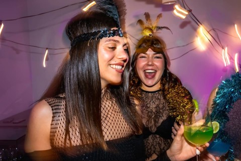 Joyful women wearing festive costumes and headbands, holding cocktails and laughing, enjoying a fun