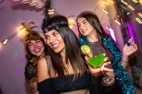 Three friends smiling and celebrating a festive costume party at home, holding a green cocktail and