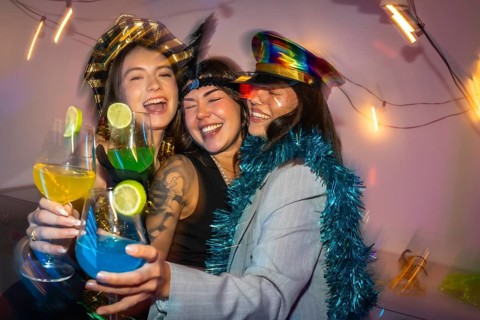 Three happy young women in festive costumes laughing and cheering with vibrant cocktails, enjoying