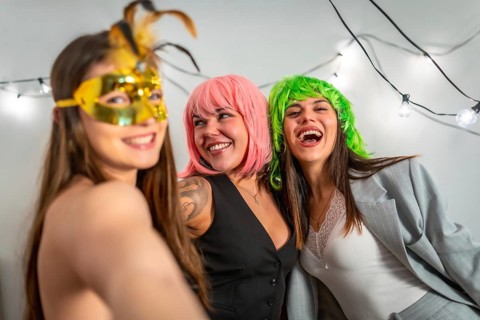 Three young adult female friends laughing and enjoying a costume party with vibrant wigs and golden
